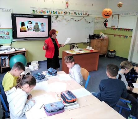 Palásthy Ágnes tanárnő felolvassa a meséjét. Mögötte az okostáblán a mesekönyvei kivetített képe látható. Az iskolás gyerekek négyes csoportokban ülnek, két összefordított pad mellett. Néhányan a feladatlapot nézegetik, mások a Tanárnőre figyelnek.